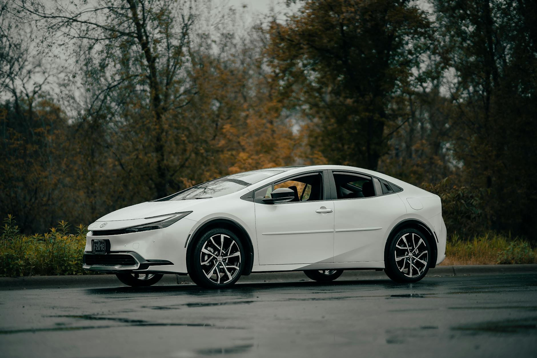 Biała hybrydowa Toyota sedan jedzie po mokrej drodze w jesiennym otoczeniu